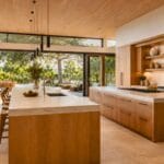 Sunlit 2026 modern kitchen with warm white oak double islands, porcelain countertops, flush-mounted appliances, and expansive glass walls opening to an outdoor patio with biophilic landscaping.